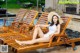 A woman in a white bathing suit and hat sitting on a wooden lounge chair.