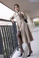 A woman in a beige trench coat and black stockings posing on a balcony.